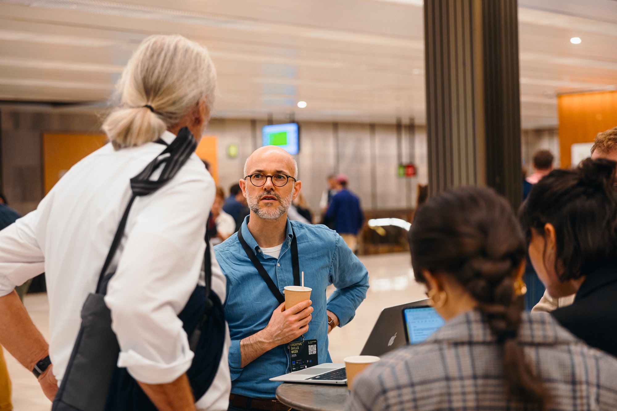 Im Vordergrund steht Henning Berthold, Projektleiter Analyse und Dialog, Innovationsnetzwerk Kreativwirtschaft der Bayern Innovativ GmbH, an einem Tisch und spricht mit einem Mann. Neben ihnen stehen zwei Frauen am Tisch, auf dem sich Laptops befinden. Im Hintergrund sind weitere Menschen in einem hellen Innenraum zu sehen.