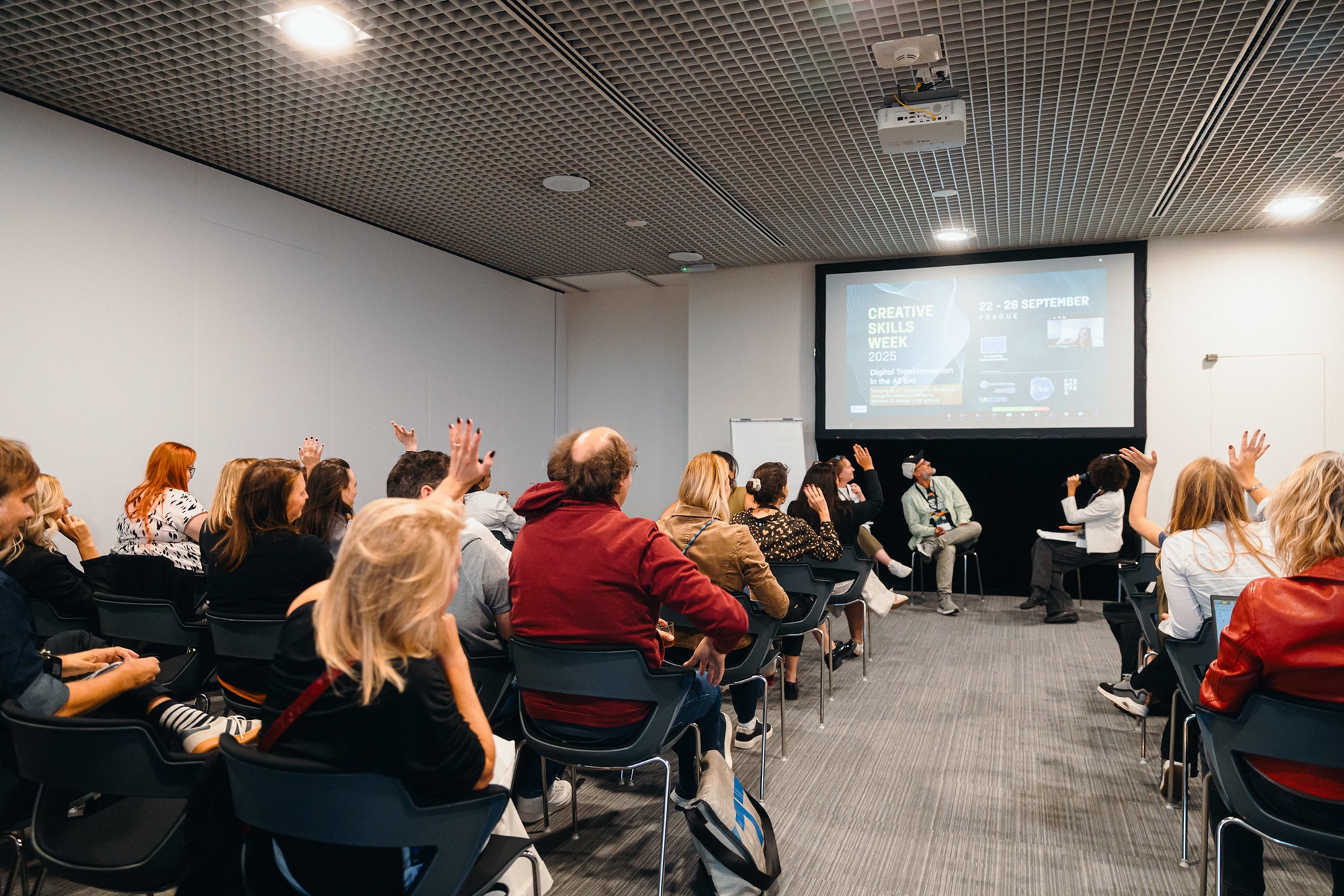 In einem Konferenzraum sitzen einige Personen auf Stühlen und nehmen an einer Veranstaltung teil. Einige heben die Hand, während vorne ein paar Personen in einer Gesprächsrunde sitzen. Auf einer Leinwand hinter ihnen ist eine Präsentation mit der Überschrift „Creative Skills Week“ zu sehen.