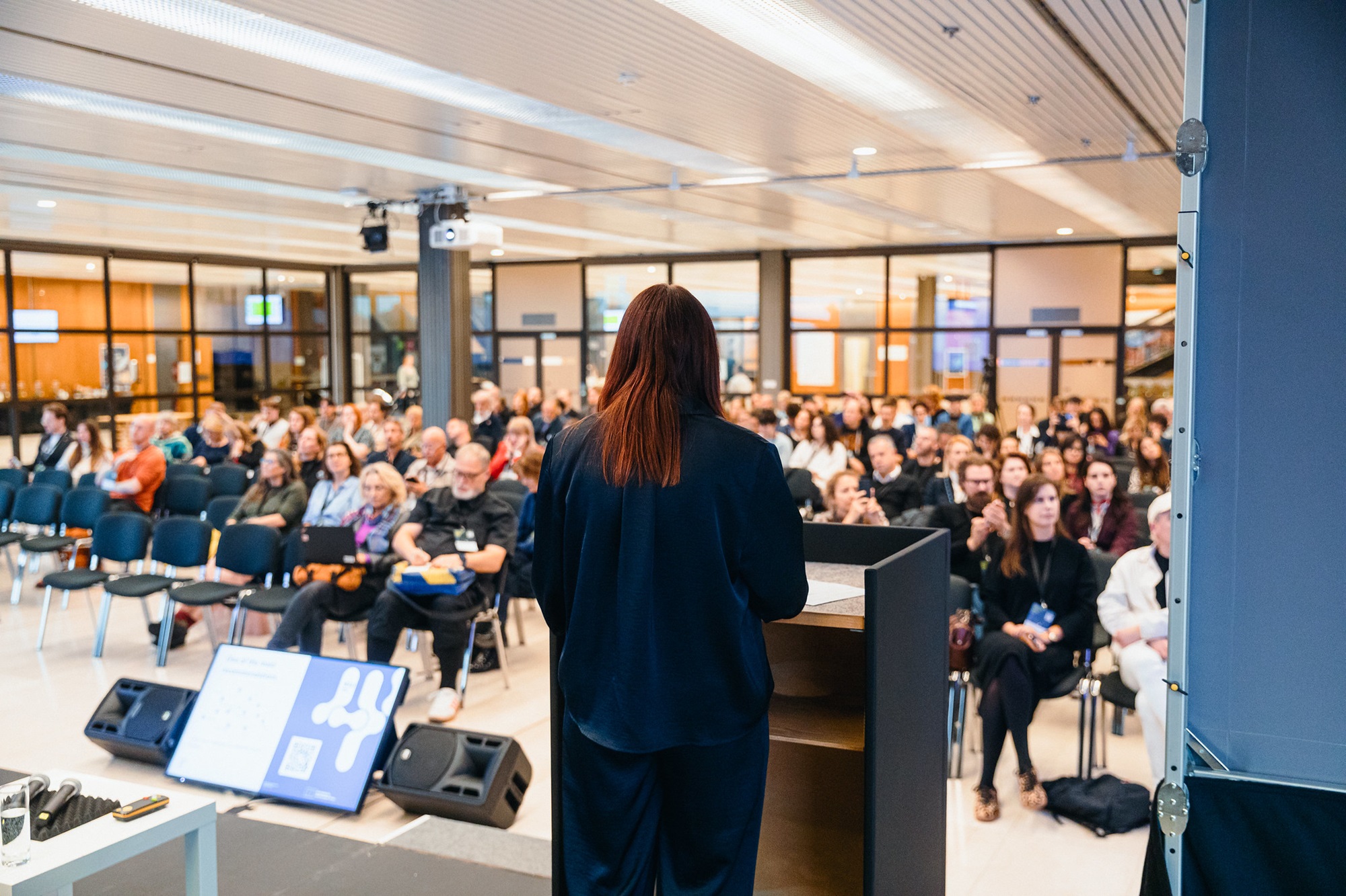 Eine Frau steht an einem Rednerpult und spricht vor einem Publikum. Sie ist von hinten zu sehen, während einige Personen auf Stuhlreihen ihr aufmerksam zuhören. Im Vordergrund stehen Lautsprecher und ein Bildschirm mit einem Veranstaltungsplan.