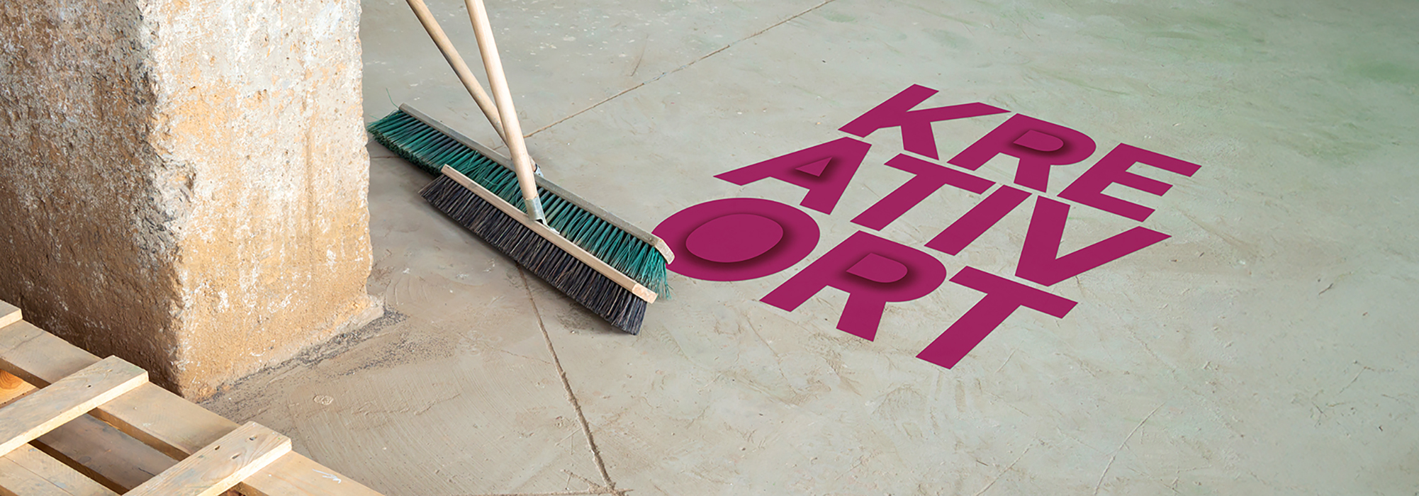 Das Bild zeigt eine glatte Betonfläche, auf der in großen, kräftigen pinken Buchstaben das Wort „KREATIVORT“ steht. Links lehnen zwei Besen an einer Wand, die Borsten berühren den Boden. Oben links ist das Logo der Auszeichnung „Staatspreis für bayerische Kreativorte“ zu sehen, bestehend aus einem stilisierten „K“ in Pink und dem Schriftzug daneben. Der Bildaufbau vermittelt den Eindruck eines vorbereiteten oder aufgeräumten Raumes, der mit Kreativität in Verbindung gebracht wird.