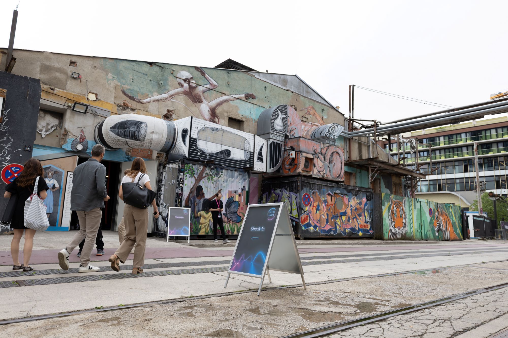 Mehrere Personen gehen an einem ehemaligen Industriegebäude mit großflächigen Graffiti entlang. Im Vordergrund weist ein Aufsteller auf den Check-in der Veranstaltung „TECH DAYS“ hin.