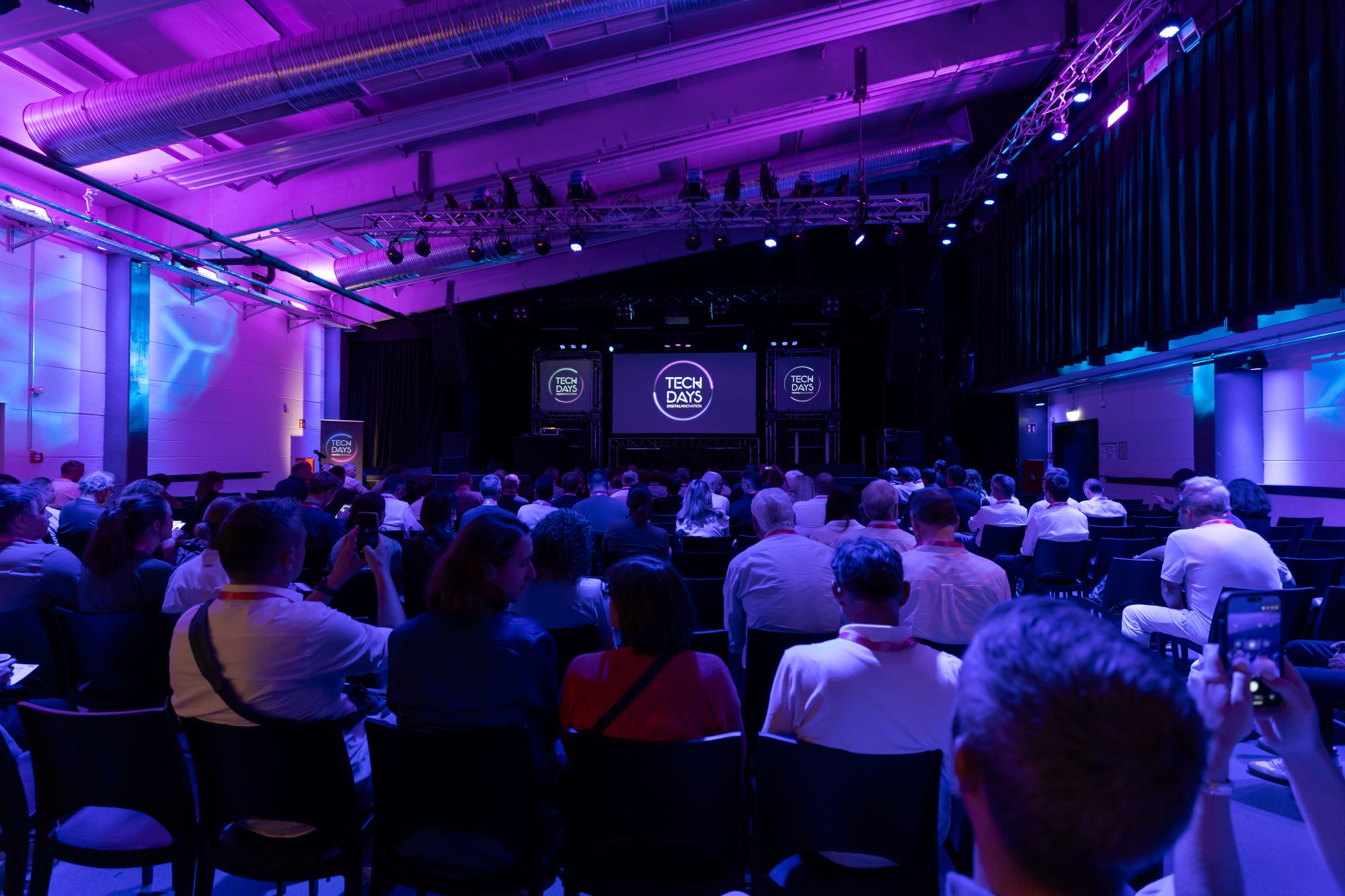 Zahlreiche Personen sitzen in einem Veranstaltungsraum und verfolgen einen Vortrag auf einer Bühne. Links und rechts der Bühne sind Bildschirme mit dem „TECH DAYS“-Logo zu sehen.