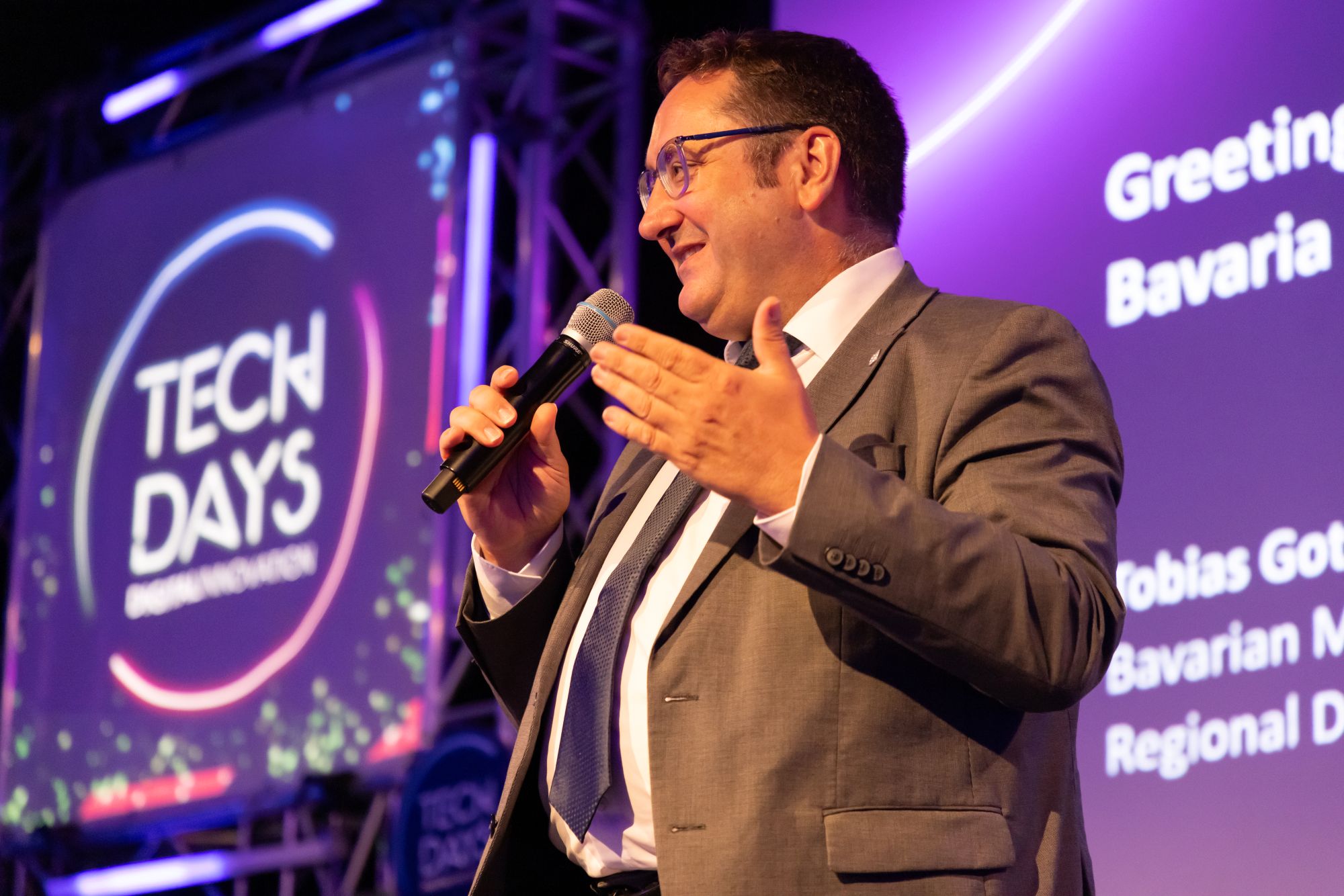 Tobias Gotthardt, Staatssekretär, Bayerisches Staatsministerium für Wirtschaft, Landes-entwicklung und Energie, steht auf einer Bühne und spricht mit einem Mikrofon. Im Hin-tergrund sind das „TECH DAYS“-Logo und eine Präsentationsfolie zu sehen.