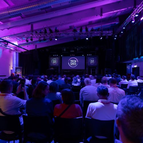 Zahlreiche Personen sitzen in einem Veranstaltungsraum und verfolgen einen Vortrag auf einer Bühne. Links und rechts der Bühne sind Bildschirme mit dem „TECH DAYS“-Logo zu sehen. 