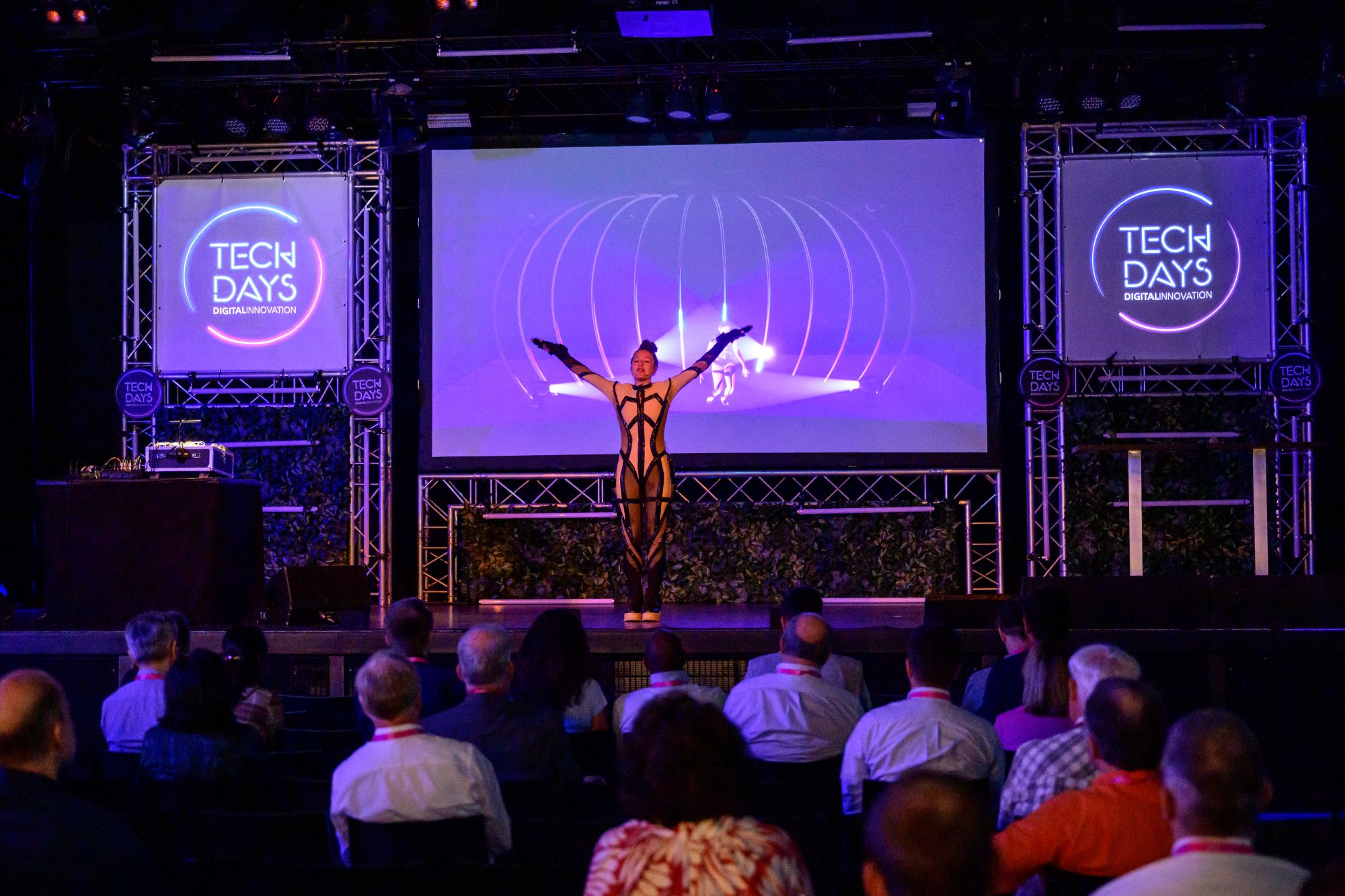 Eine Tänzerin steht mit ausgestreckten Armen auf einer Bühne und führt vor dem Publikum eine Performance auf. Hinter ihr ist eine großflächige Projektion und beleuchtete „TECH DAYS“-Logos zu sehen.