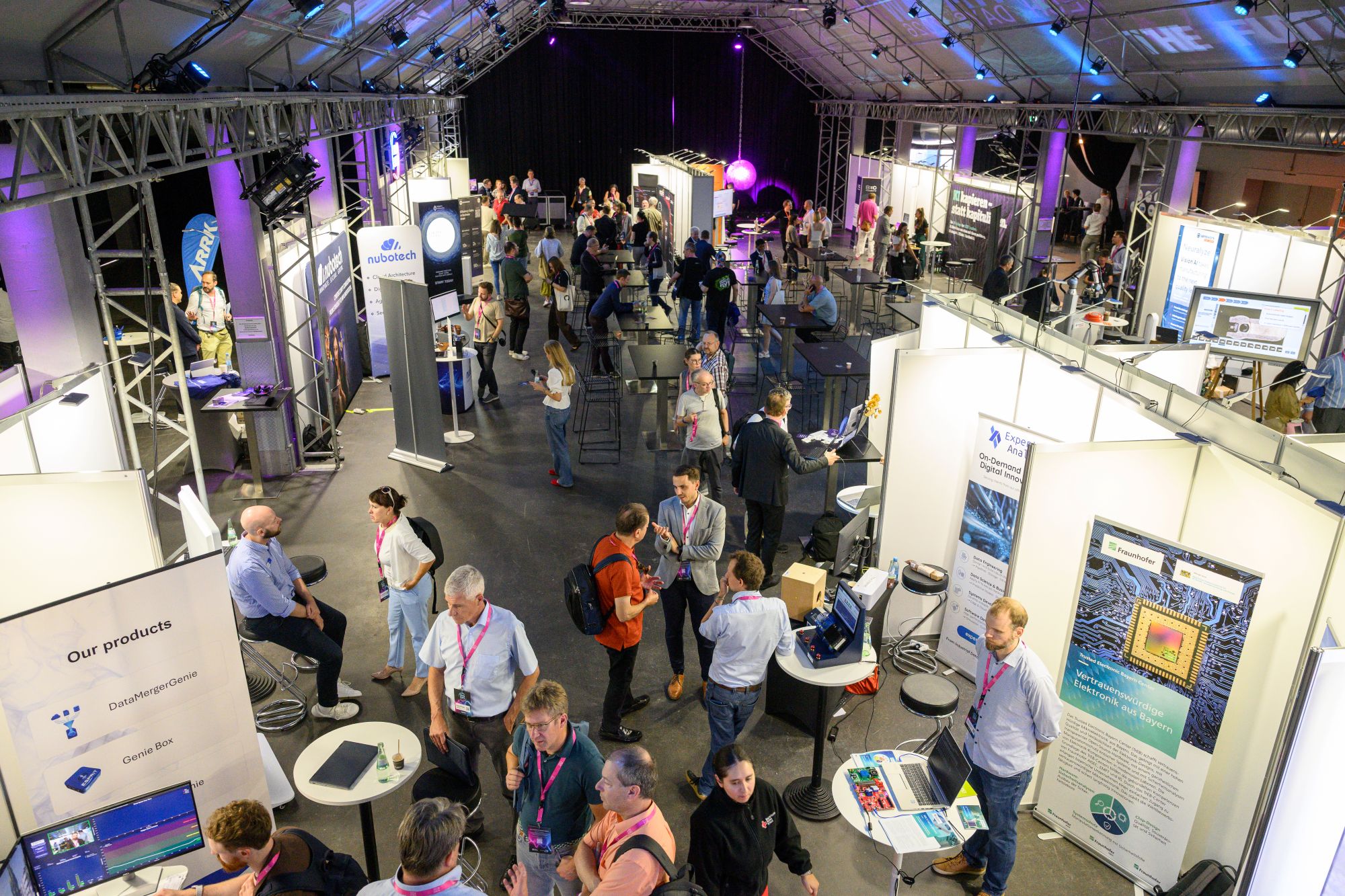 Der Blick von oben zeigt eine Ausstellung mit zahlreichen Messe-ständen. Besucherinnen und Besucher bewegen sich zwischen den Ständen und führen Gespräche.