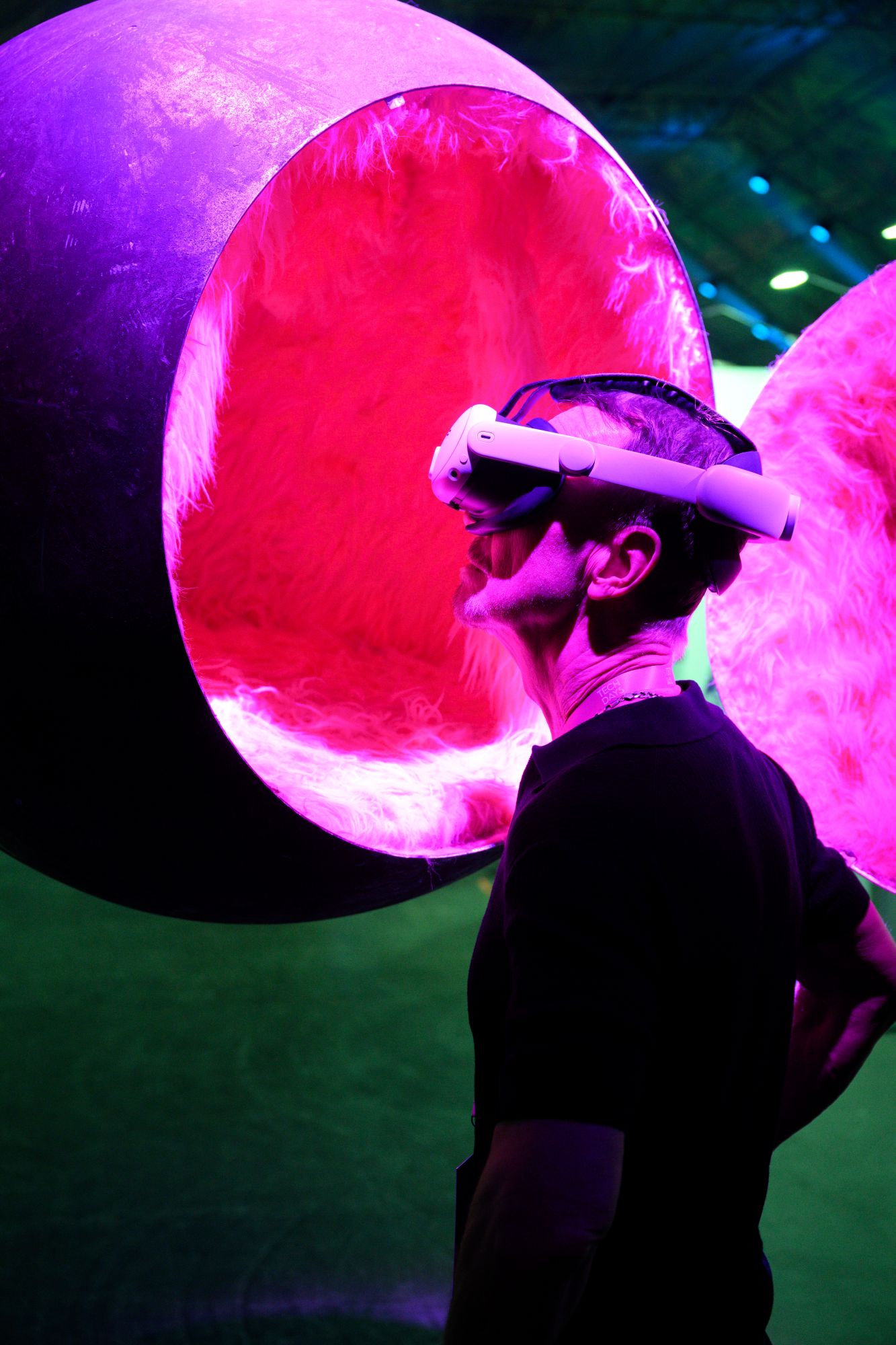 Eine Person trägt eine Virtual-Reality-Brille und blickt nach oben. Im Hintergrund sind große, leuchtende kugelförmige Installationen in Pink- und Violetttönen zu sehen.