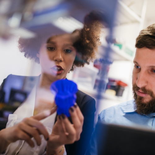ChatGPT:  Das Bild zeigt zwei Personen in einem Labor oder Technikum, die gemeinsam ein komplex geformtes, blaues 3D-Druckobjekt begutachten.