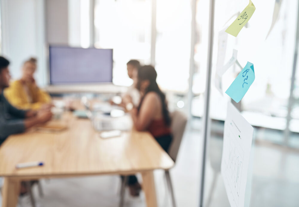 Personen sitzen an einem Tisch zusammen, im Vordergrund sind Post-its an einer Glaswand zu sehen