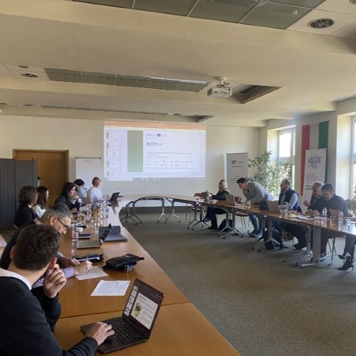 Menschen sitzen an einem langen Tisch in einem Raum und verfolgen eine Präsentation