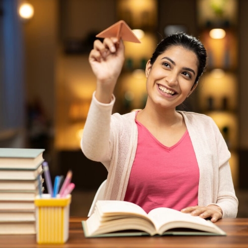 Junge Frau sitzt an einem Schreibtisch mit aufgeschlagenem Buch, hält lächelnd ein Papierflugzeug in die Luft – neben ihr ein Stapel Bücher und ein Stiftebecher.