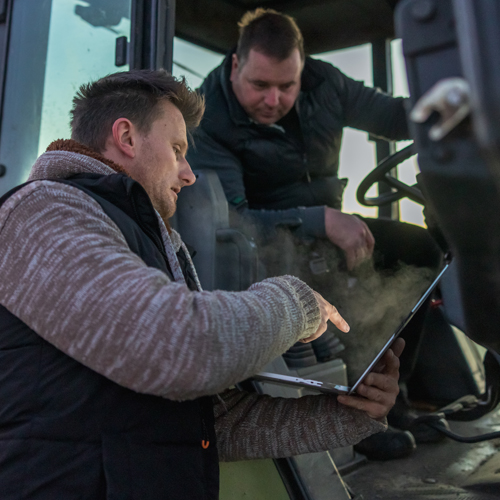 Zwei Männer nutzen einen Laptop, während sie in einem Traktor sitzen.