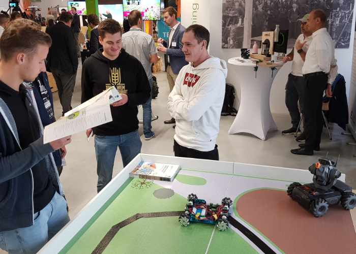 In der Ausstellung stehen an einem Stand 3 Männer im Gespräch vertieft