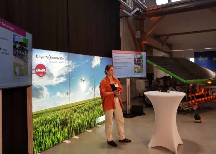 Ein Referent auf der Bühne mit Stehtisch und im Hintergrund eine Motivwand auf der ein Acker mit weißblauem Himmel abgebildet ist