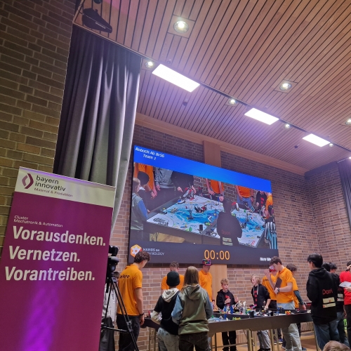 Im Vordergrund steht eine Leinwand mit dem Logo und den Farben des Clusters Mechatronik & Automation. Im Hintergrund präsentieren Kinder ihre Projekte an einem großen Tisch, während ihre Arbeiten gleichzeitig auf einem großen Bildschirm an der Wand übertragen werden