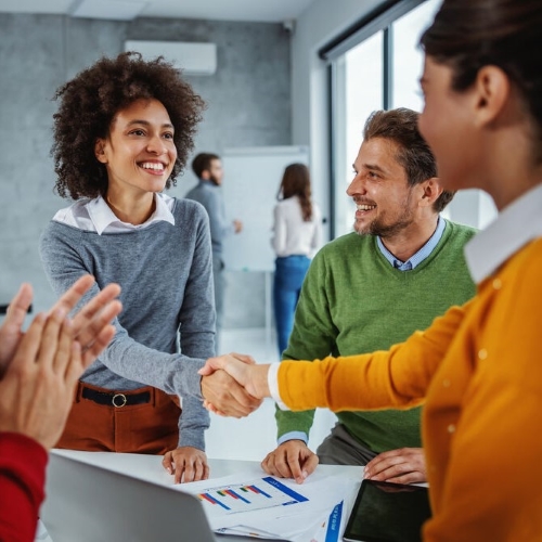 Eine Frau inmitten einer Gruppe von Menschen bei einer Fachveranstaltung lächelt freundlich, während sie einer anderen Frau zur Begrüßung die Hand schüttelt.