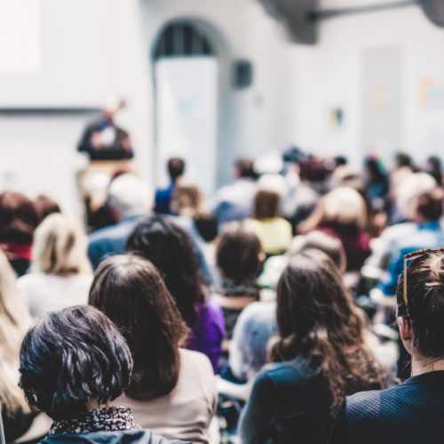 Publikum aus mehreren Menschen von hinten zu sehen vor einem Vortragenden