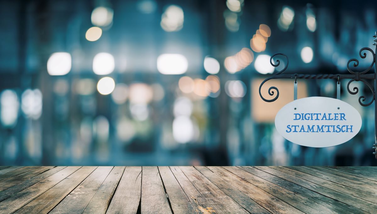 Das Bild zeigt eine rustikale Holzoberfläche im Vordergrund, die als Tisch oder Theke interpretiert werden kann. Im Hintergrund ist eine unscharfe Szene mit Lichtern zu sehen, die eine Bar oder ein Restaurant andeuten könnte. Rechts hängt an einem kunstvoll geschmiedeten Schildhalter ein ovales Schild mit der Aufschrift "DIGITALER STAMMTISCH" in blauer Schrift.