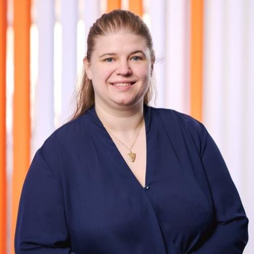 Person mit zusammengebundenem Haar trägt eine dunkelblaue Bluse und eine Halskette mit einem herzförmigen Anhänger vor einem Hintergrund mit orange-weißen vertikalen Streifen