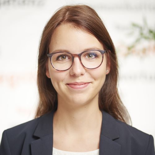 Das Bild zeigt eine Person in einem weißen Hemd mit Kragen und einem dunklen Blazer vor einem schlichten, hellen Hintergrund.