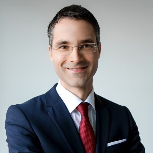 Das Bild zeigt ein Porträt eines Mannes mittleren Alters mit kurz geschnittenem, dunkelbraunem Haar. Er trägt eine dunkelblaue Anzugjacke, ein weißes Hemd und eine dunkelrote Krawatte. Er trägt eine schlichte, silberne Brille mit dünnen Bügeln. Der Mann lächelt leicht und schaut direkt in die Kamera. Der Hintergrund ist einfarbig hellgrau.