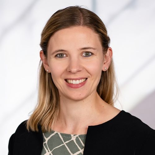 Das Bild zeigt eine junge Frau mit schulterlangen, blonden Haaren. Sie trägt ein schwarzes Sakko über einem grün-beige gemusterten Oberteil. Sie lächelt freundlich in die Kamera. Der Hintergrund ist unscharf und hell, mit einem Muster, das an Licht und Schatten erinnert. Das Foto wirkt professionell und könnte ein Bewerbungsfoto oder ein Porträt für eine Webseite sein.