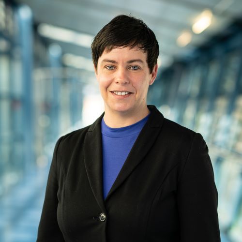 Das Bild zeigt eine Frau im schwarzen Blazer, darunter ein blaues TShirt. der Hintergrund ist verschwommen