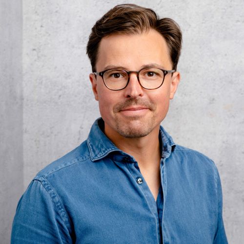 Das Bild zeigt ein Porträt eines Mannes mittleren Alters mit braunen Haaren und einer Brille. Er trägt ein hellblaues Jeanshemd und blickt direkt in die Kamera. 