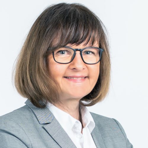 Das Bild zeigt ein Porträt einer Frau mittleren Alters. Sie hat kurzes, braunes Haar mit Pony und trägt eine schwarze Brille. Sie lächelt freundlich in die Kamera. Bekleidet ist sie mit einer weißen Bluse und einem hellgrauen Blazer.