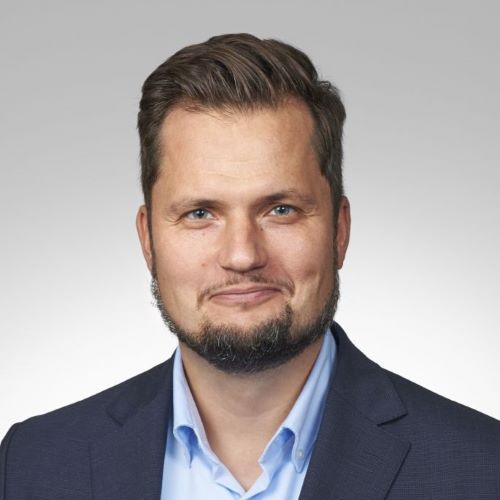 Ein Mann mit einem Bart trägt ein blaues Hemd und ein schwarzes Sakko. Er hat braune Haare und lächelt.