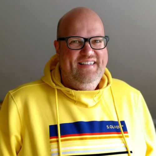 Auf dem Bild ist ein Herr mit Glatze zu sehen und schwarzer Brille. Er trägt einen gelben Hoodie