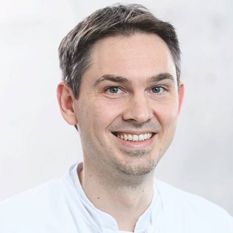 Auf dem Bild ist ein Herr mit einem weißen Hemd zu sehen. Er lacht in die Kamera