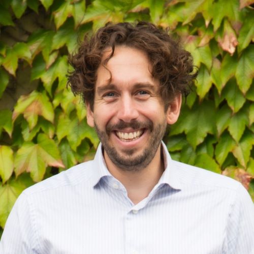 Das Bild zeigt einen Mann mit lockigem Haar und einem Bart, der lächelt. Er trägt ein hellblaues Hemd. Im Hintergrund befindet sich eine Wand, die mit Efeu bewachsen ist.