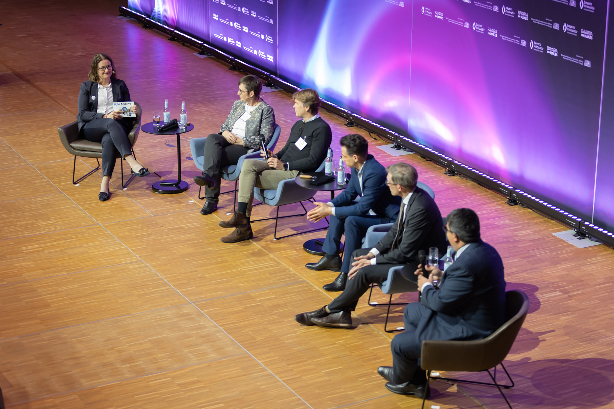 Weitwinkelaufnahme einer Podiumsdiskussion auf der Bühne vor Publikum. Das Bild entstand beim AM Kongress.