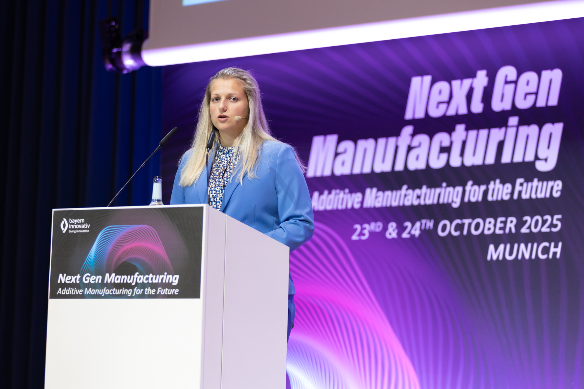 Frau spricht am Rednerpult mit der Aufschrift „Next Gen Manufacturing – AM Kongress München 2025“. Entstanden beim AM Kongress.