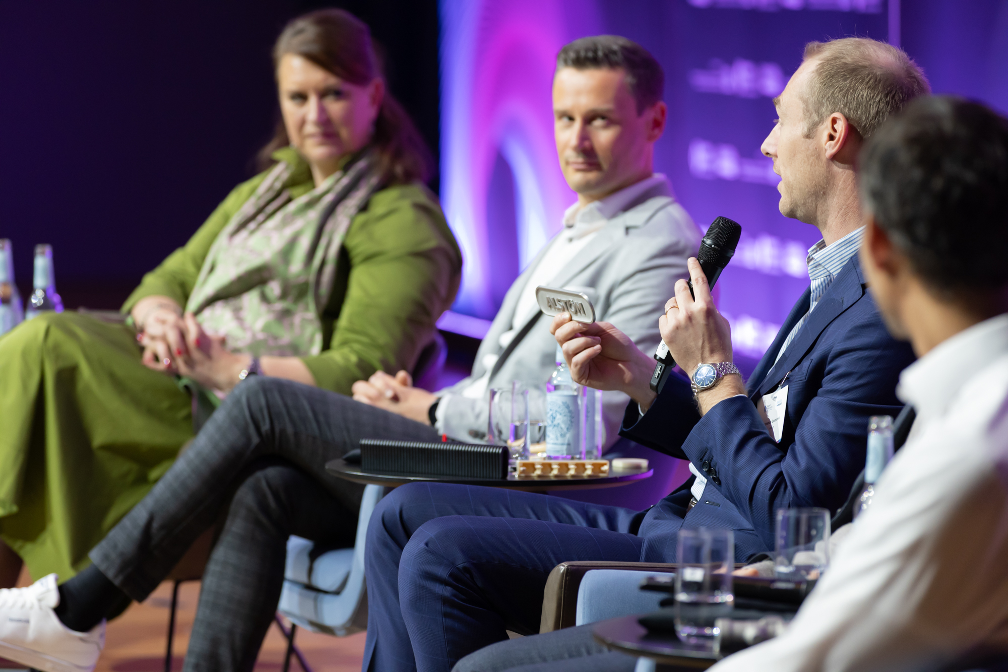 Drei Personen sitzen auf einer Bühne in Gesprächsrunde, vor lilafarbenem Hintergrund. Das Foto entstand beim AM Kongress.