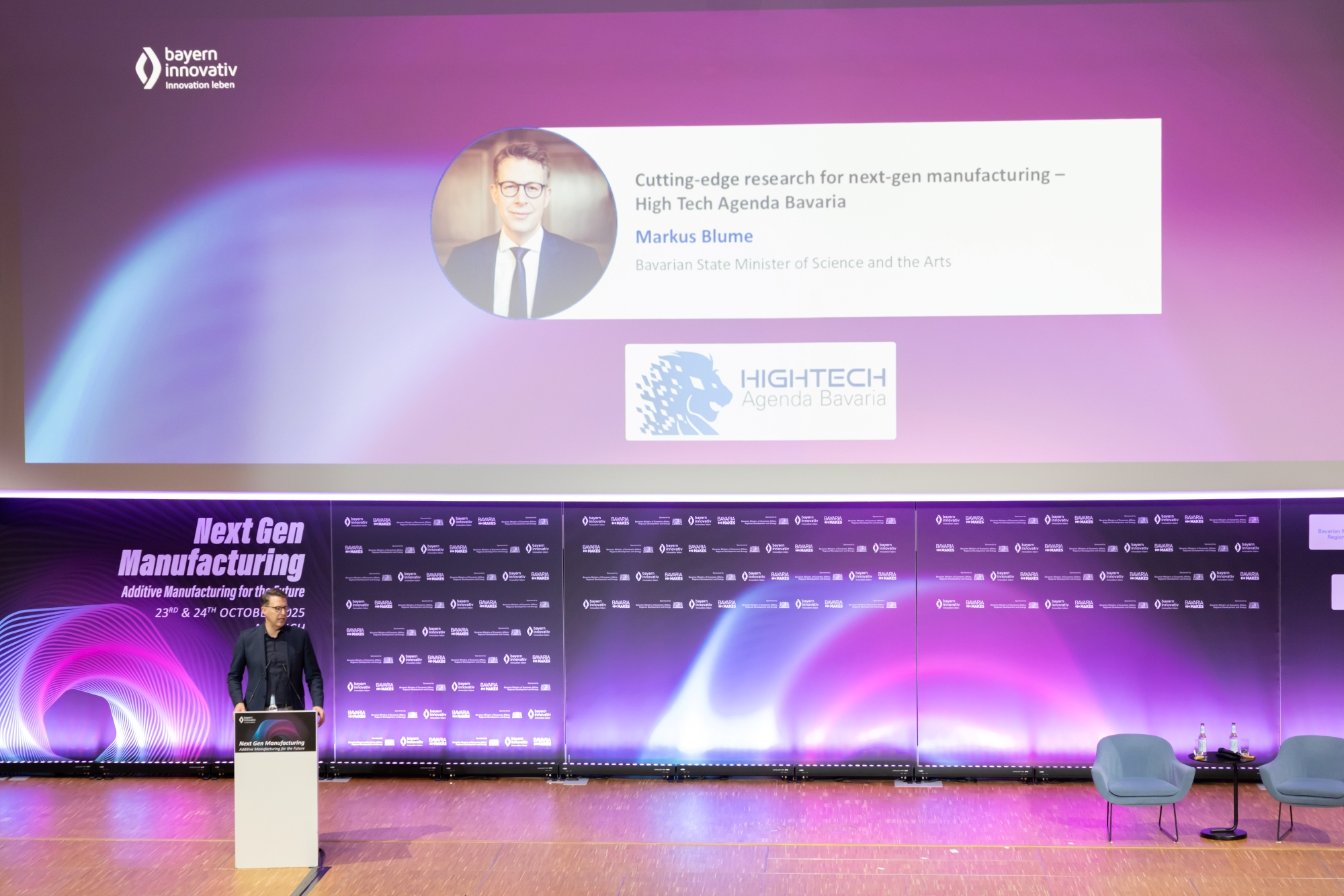 Große Leinwand mit Titel und Sprechername des AM Kongresses, Bühne in lila Beleuchtung.