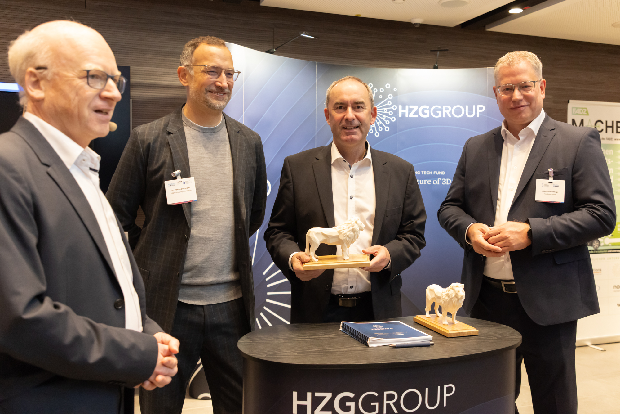 Mehrere Männer stehen im Gespräch an einem Messestand, einer hält eine Trophäe. Aufgenommen beim AM Kongress.