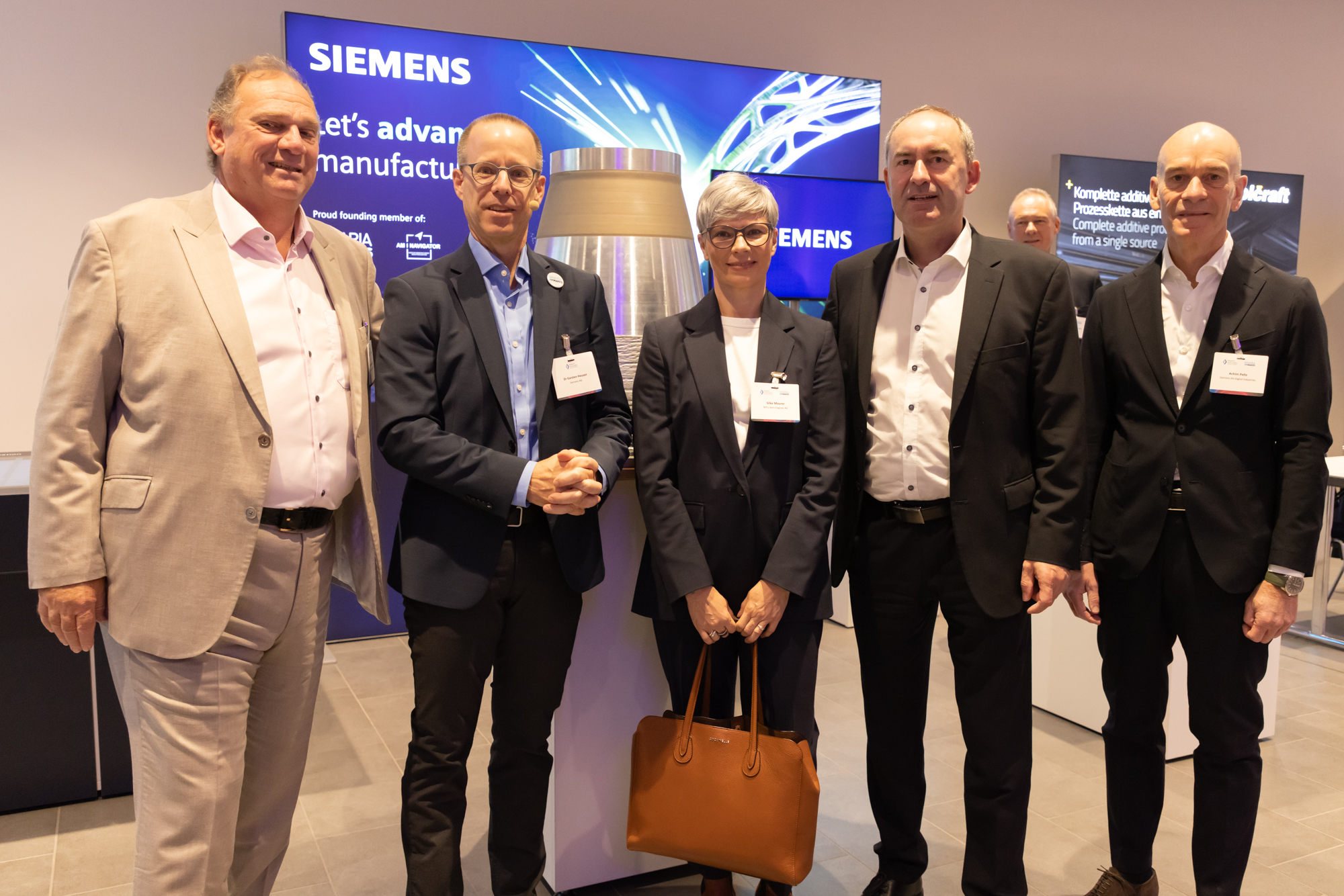 Vier Männer posieren gemeinsam vor einem Messestand mit Firmenlogos. Entstanden beim AM Kongress.