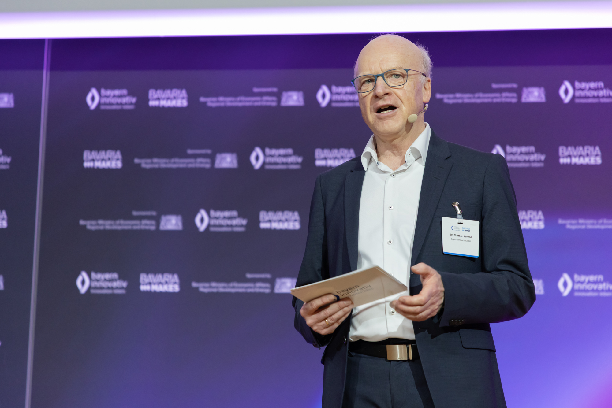 Moderator oder Redner spricht auf der Bühne, hinter ihm eine violett beleuchtete Wand mit Sponsorenlogos. Das Bild stammt vom AM Kongress.