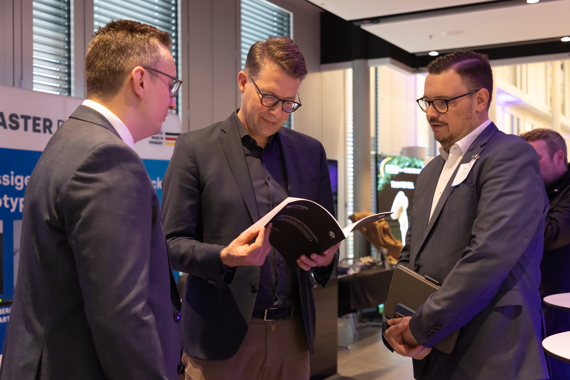 Drei Männer unterhalten sich in einer Ausstellung oder Networking-Area, einer zeigt etwas in einem Heft. Foto vom AM Kongress.
