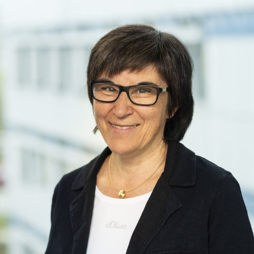 Frau mit Schwarzer Brille und schwarzen Haaren in weißem Tshirt, Kette und schwarzem Blazer