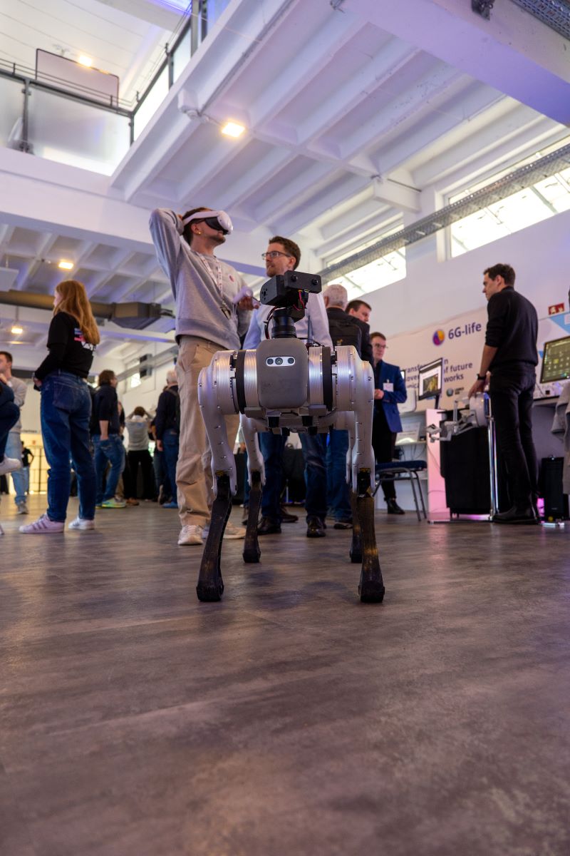 A view into the exhibition, showing a man with augmented reality glasses on his head and a 4-leged robot in front.