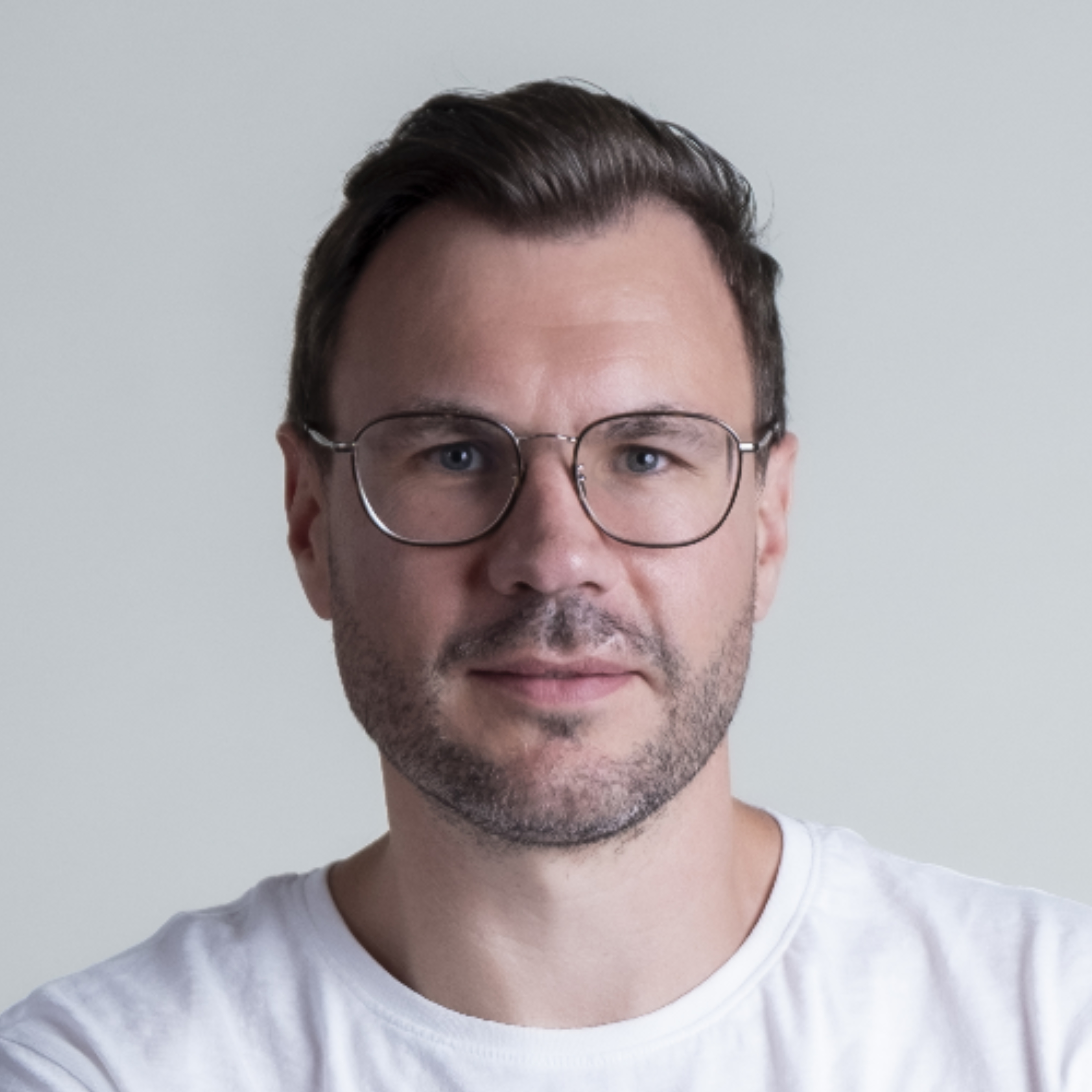 Das Bild zeigt Daniel De Monte der wemonte AG, einen Mann mit dunkelbraunen Haaren, dunkler Brille mit Metallgestell und einem frei Tage Bart, der ein weiße T-Shirt trägt. Der Hintergrund ist hellgrau