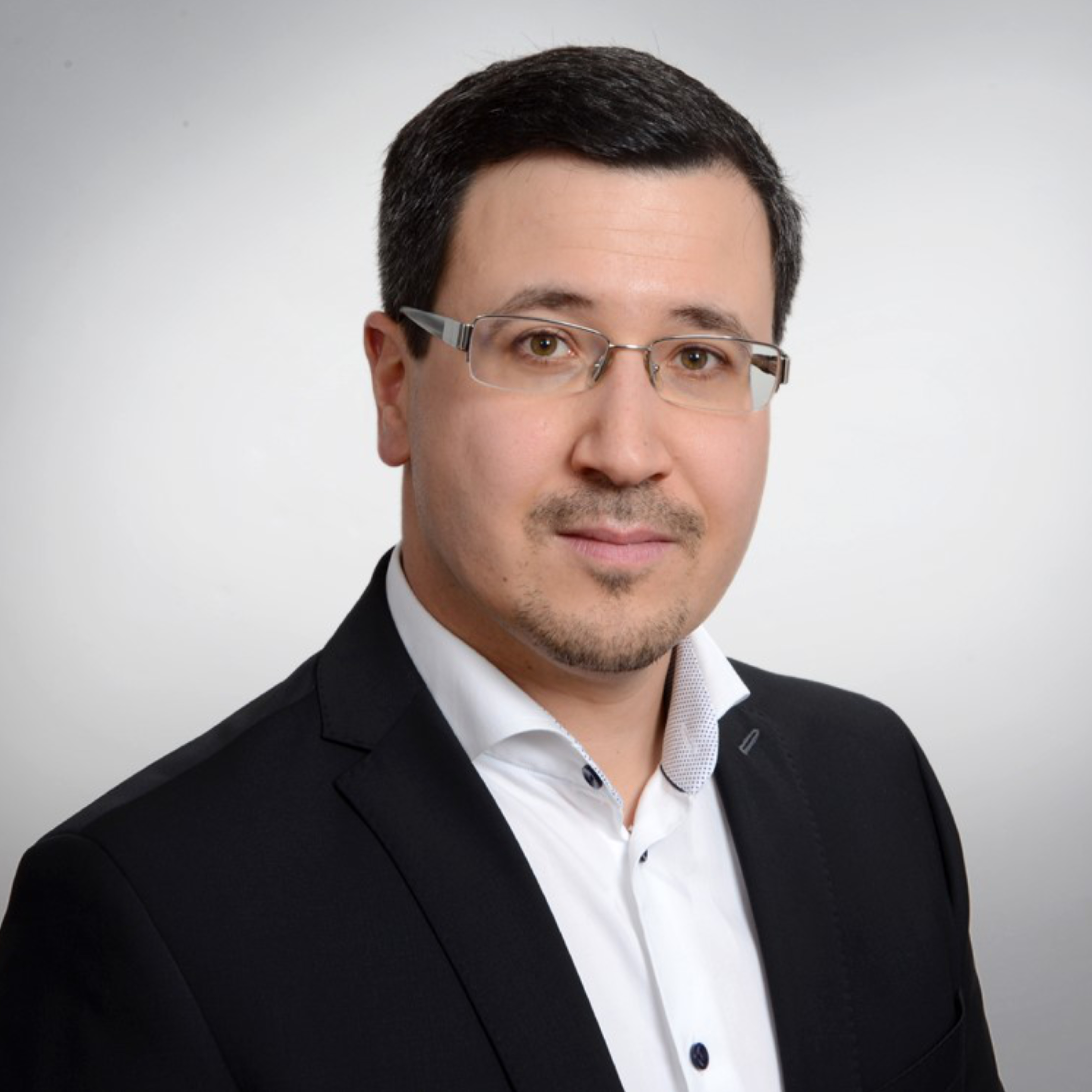 Portraitbild von Dr. Frédéric Holzmann, einem schwarzhaarigen Mann mit Brille, weißem Hemd und schwarzem Sakko