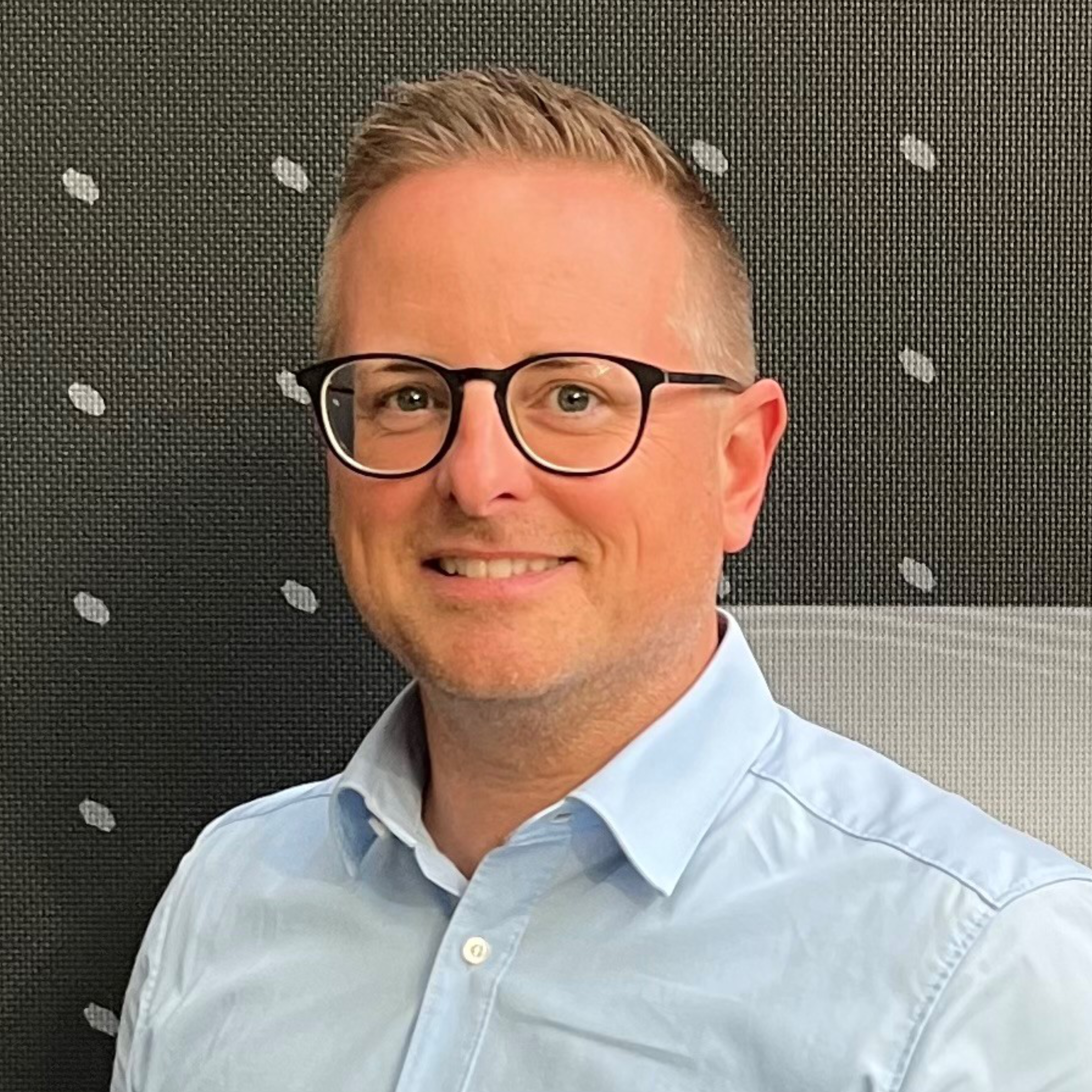 Portraitbild des Referenten Norbert Zundl, welches einen lächelnden Mann mit runder schwarzer Brille in hellblauem Hemd und kurzen blonden Haaren vor einem dunkelgrauen Hintergrund mit weißen Punkten zeigt.