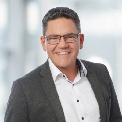 Bild von Oliver Druhm der Dräxlmaier Group, welches ihn in einem weißen Hemd mit grau-braunem Sakko vor einem grauen Hintergrund zeigt. Er hat braune Haare, lächelt und trägt eine eckige Brille mit Metallgestell