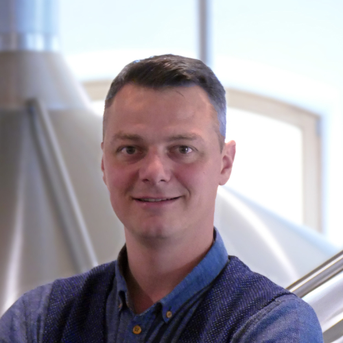 Ein Mann in formeller Kleidung steht vor einem Sudkessel.