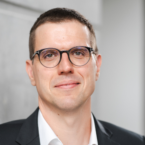 Ein Mann in formeller Kleidung steht vor einer Wand.