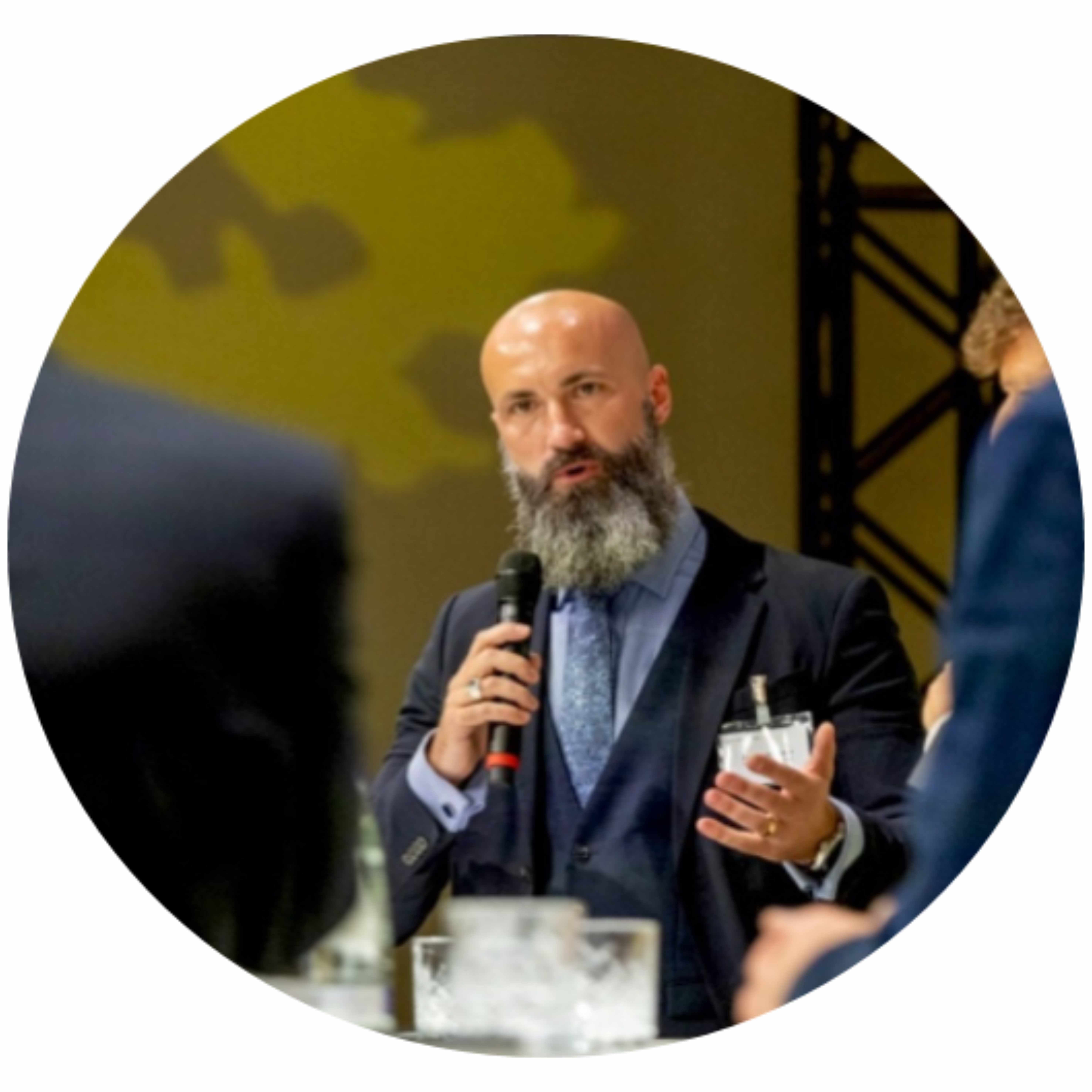 Bild von Christoph Müller, einem Mann ohne Haare und einem langen grauen Bart, der ein Mikrofon in der Hand hält und ein dunkelblaues Sakko mit einer dunkelblauen Weste und einem hellblauen Schal trägt.